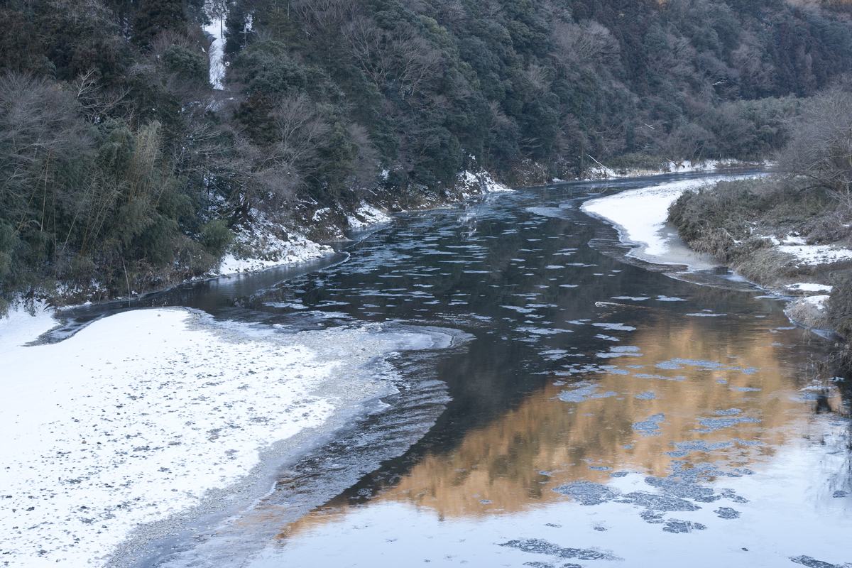 久慈川河面漂著一片片淺色碎冰往下游流,左岸整片結成白色冰灘,兩側山坡是覆雪的落葉林