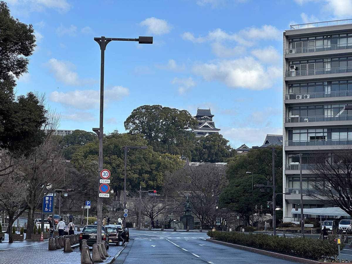 熊本市區街道往北看:雙線道路筆直通向一片大樹林,樹林上方露出熊本城天守,路中間有一座銅像,左邊是計程車招呼站與寫著「シアーズホーム夢ホール」的藍色看板,右邊是一棟灰色辦公大樓,藍天白雲