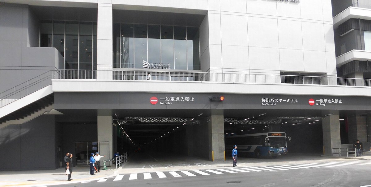熊本桜町巴士總站的巴士出入口:灰色大樓一樓有兩個巴士進出的開口,上方黑色橫條寫著「桜町バスターミナル Bus Terminal」與兩個「一般車進入禁止」,二樓玻璃牆上有「熊本城ホール」字樣,右邊開口裡停著一輛藍白色巴士