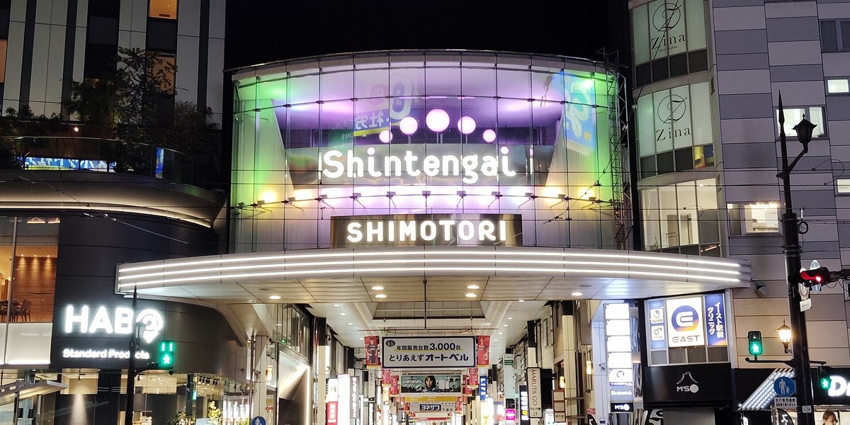 夜晚的下通拱廊入口:弧形玻璃帷幕上亮著白字「Shintengai」,下方招牌寫「SHIMOTORI」,入口雨棚打著白光,往裡看是一條掛滿店家招牌的長拱廊,左邊大樓有「HAB」字樣