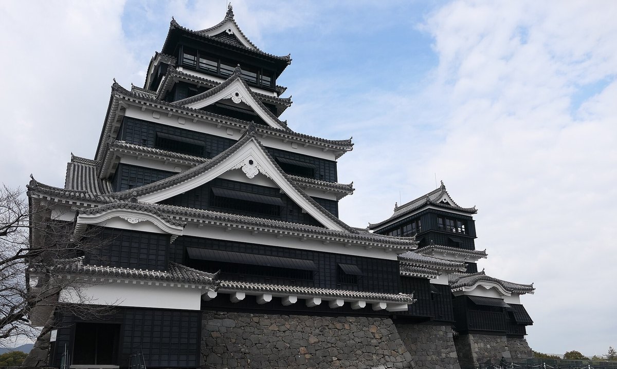 從斜前方仰拍熊本城大天守:黑色板牆、白色破風與多層灰瓦屋簷,下半部是灰色石垣,右邊接著較矮的小天守與一段玻璃扶手的參觀步道,藍天白雲