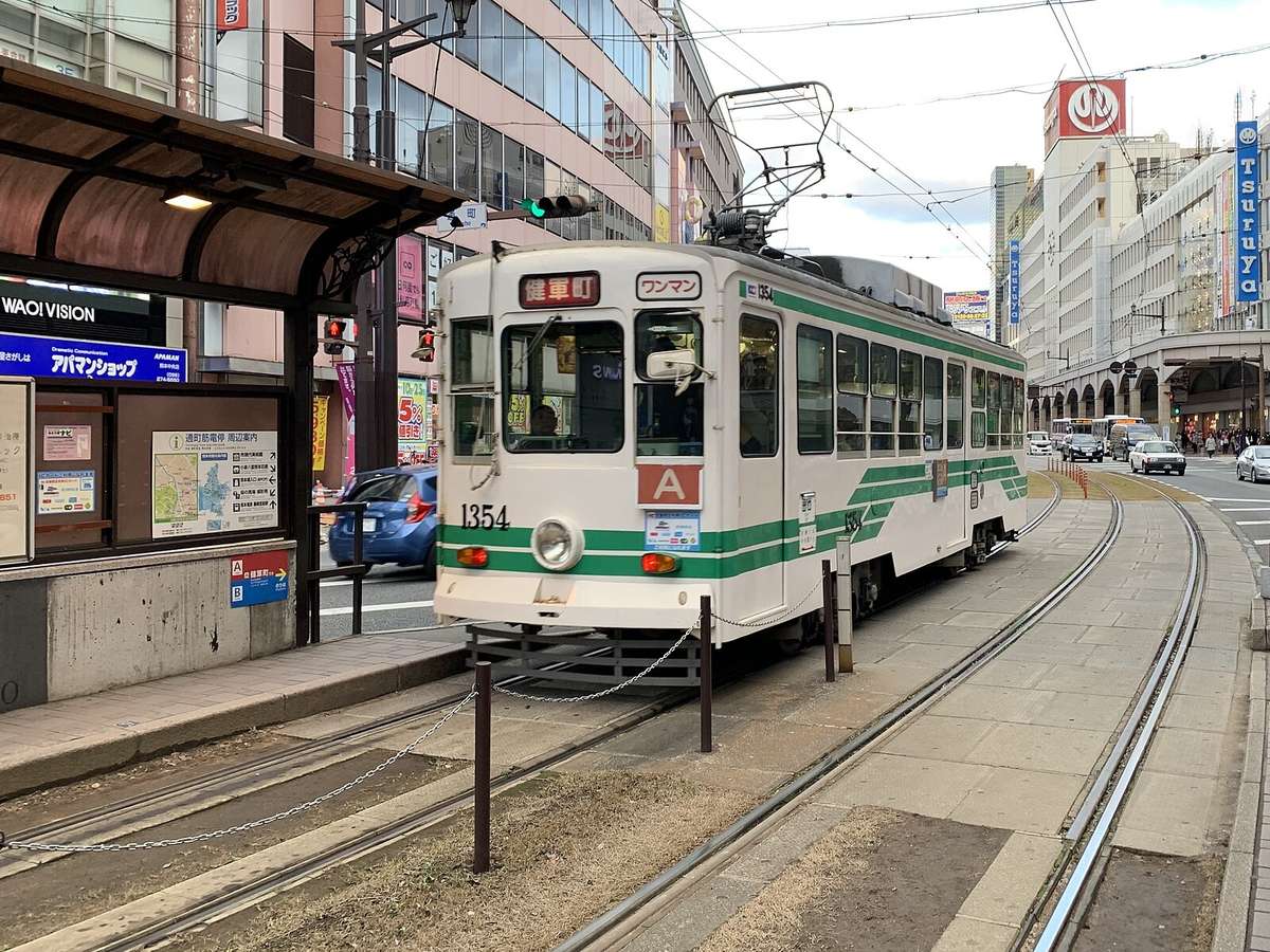 熊本市電通町筋電停:一輛白底綠條紋、車頭寫著「健軍町」的單節路面電車停在月台旁,左邊電停的看板寫「通町筋電停 周辺案内」,右後方是印著 Tsuruya 的鶴屋百貨大樓與騎樓