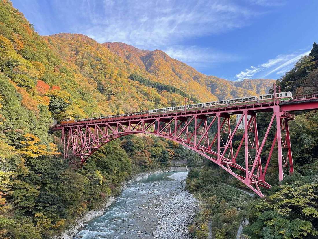 Autumn in the Kurobe Gorge: a torokko train crossing a red steel arch bridge, a pale stony river below and the far hillside solid orange and yellow