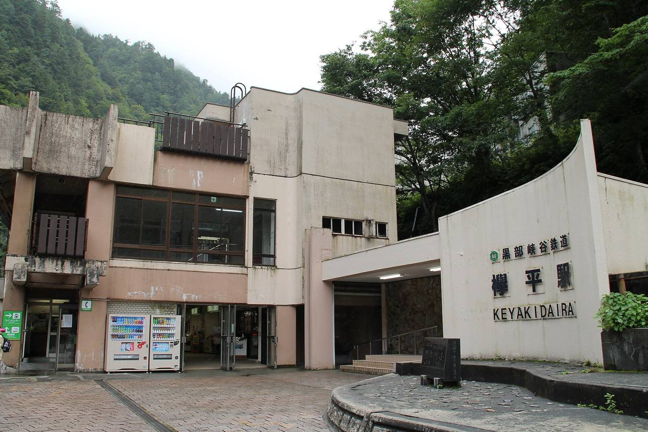 The forecourt of Keyakidaira station: a curved white wall carrying the station name on the right, the concrete station building with vending machines and a kiosk on the left, and a covered ramp to the ticket gates between them