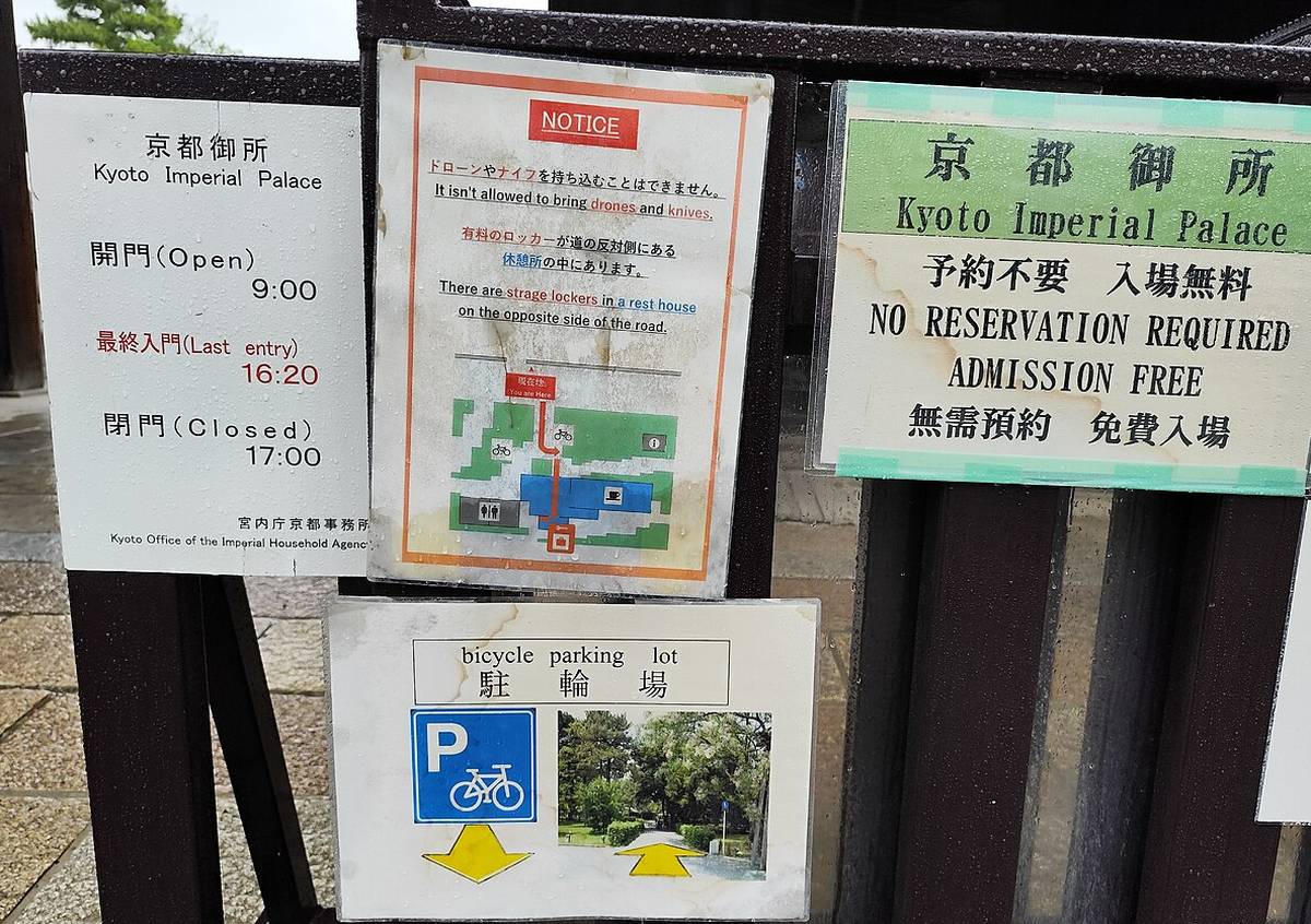 京都御所入口柵欄上的幾張告示：綠底牌子寫「京都御所 予約不要 入場無料」與中文「無需預約 免費入場」，左邊白牌寫開門 9:00、最終入門 16:20、閉門 17:00，中間是禁帶無人機與刀具的告示