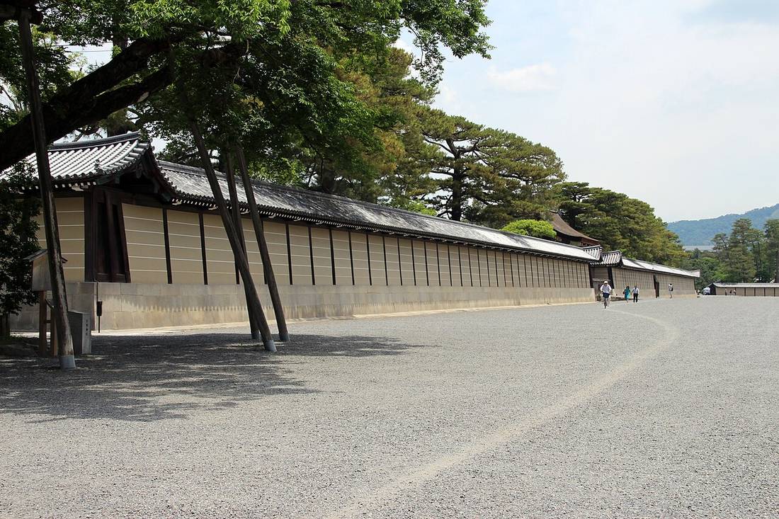 京都御苑內寬闊的碎石路，左側是京都御所米黃色的築地塀與瓦頂，牆後是高大的松樹，遠處有人騎腳踏車與步行