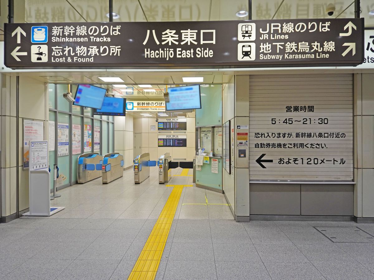JR 京都站新幹線八條東口剪票口外側：頭頂深咖啡色吊牌正中寫「八条東口 Hachijō East Side」，兩側箭頭分別指向新幹線のりば、失物招領處、JR 線のりば與地下鐵烏丸線；後方是一排藍色新幹線剪票機，右邊是拉下鐵捲門的售票處