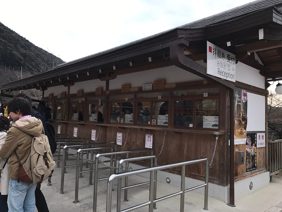 The ticket office of a Kyoto temple: seven wooden windows in a row under a tiled eave, a white sign overhead reading haikan-ken uketsuke with Korean and the word Reception, hand-lettered price boards above the windows, printed fares taped below them, and stainless steel rails marking out a queue