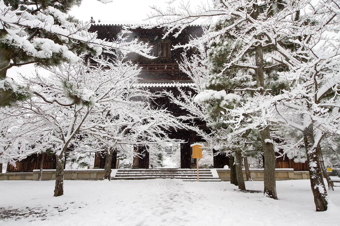 A two-storey temple gate in heavy snowfall, its roof and the surrounding pines and cherry trees loaded with snow, the stone steps and the whole forecourt covered except for a single line of footprints, a wooden noticeboard standing beside the gate, and nobody in sight