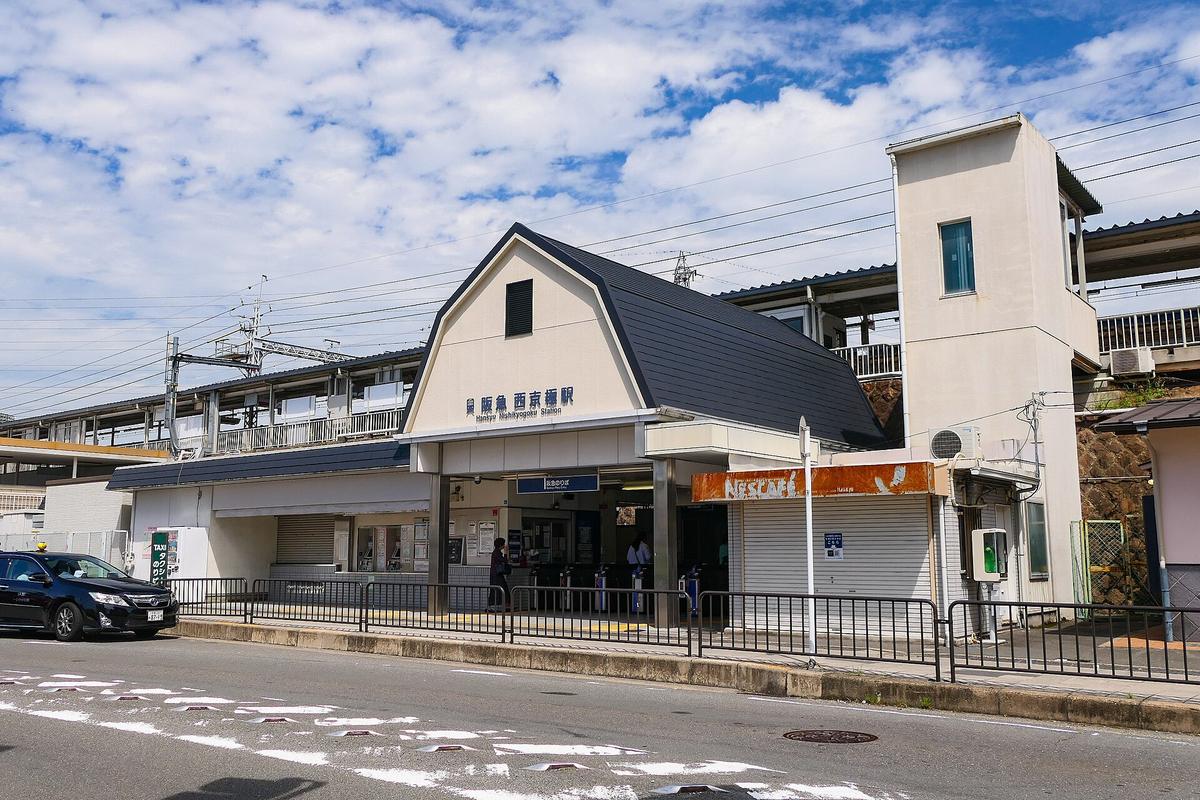 阪急西京極站的地面站房,米白色山形屋頂上寫著「阪急 西京極駅」,下方是自動改札口,左邊是計程車招呼站,後方高架上是月台