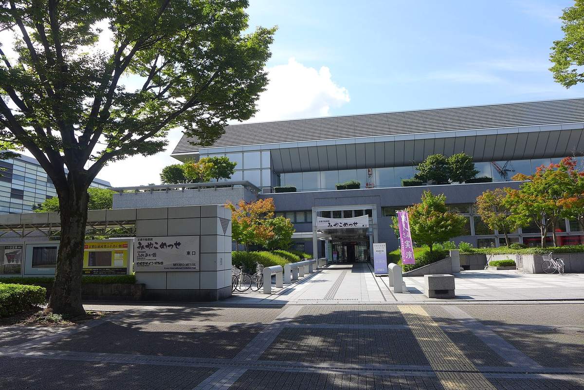 みやこめっせ東口:左邊石牆招牌寫著「京都市勧業館 みやこめっせ 東口 MIYAKO MESSE」,正面玻璃入口上方掛著みやこめっせ的布幕,前面是石板步道與腳踏車停車處