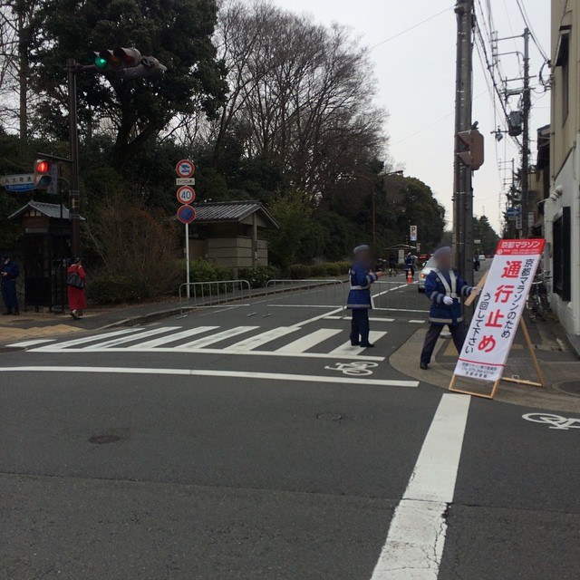 路口立著白底紅字的直立看板,上方紅框寫「京都マラソン」,大字「通行止め」,旁邊寫「京都マラソンのため う回して下さい」,兩名穿藍色制服的交通指揮人員站在斑馬線旁,後方路口用柵欄封住