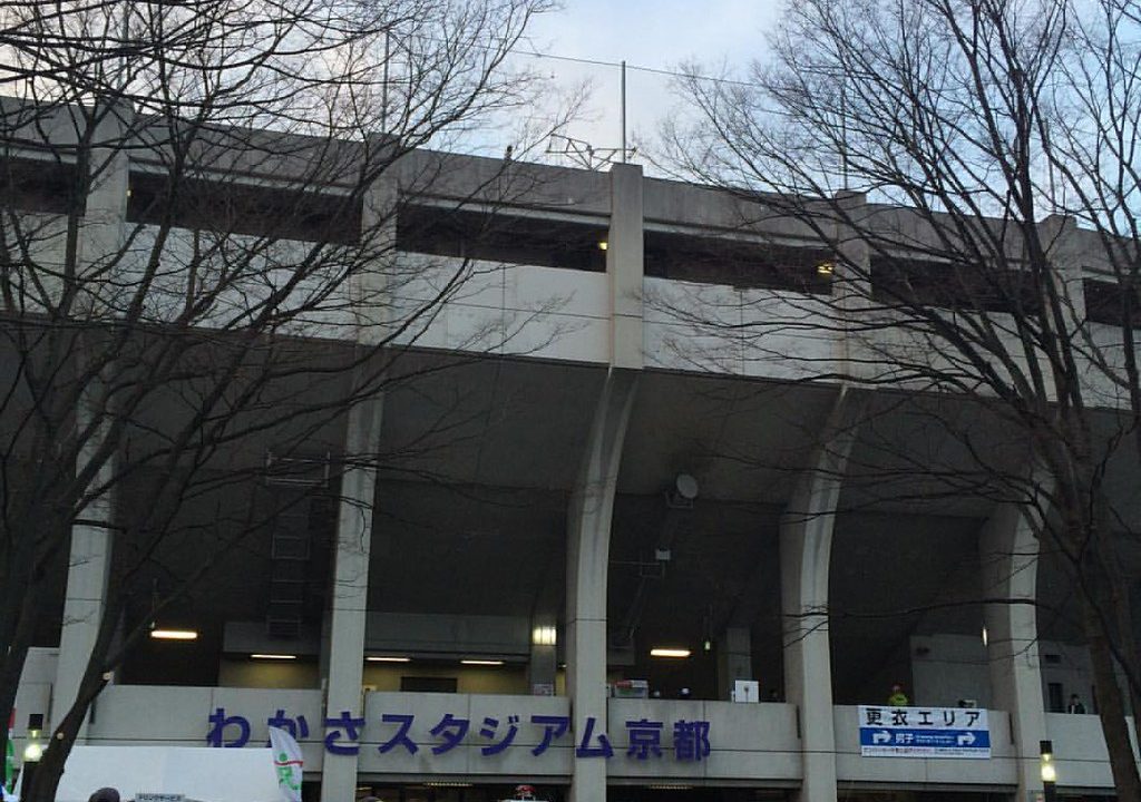 比賽日清晨的西京極田徑場外觀,灰色混凝土看台下方大字寫著「わかさスタジアム京都」,右側掛著「更衣エリア 男子」的藍白指示牌,前景是冬天的枯樹枝