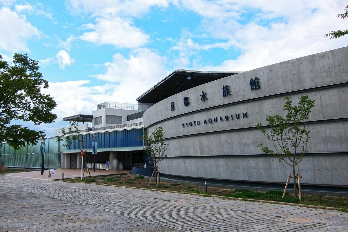 京都水族館外觀:弧形的清水混凝土外牆上寫著「京都水族館 KYOTO AQUARIUM」,前方是石板步道與剛種下的小樹,左邊是玻璃外牆的入口區,遠處有兩位行人(臉部已模糊)