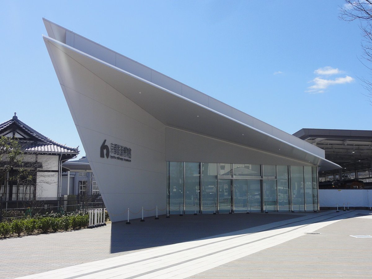 京都鐵道博物館入口大廳外觀:白色的斜屋頂像機翼一樣往左上翹起,牆上有「京都鉄道博物館 kyoto railway museum」字樣與館徽,正面是整片玻璃門,左後方是一棟和風木造建築,晴天無人