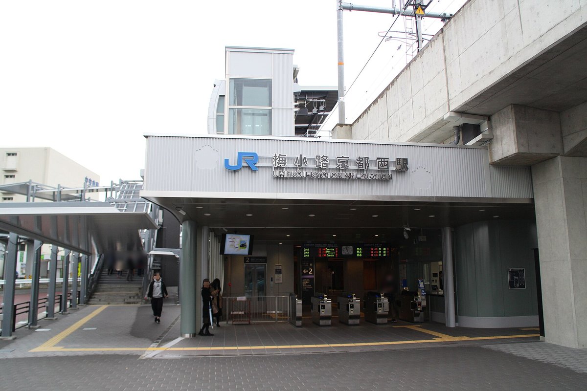 JR 梅小路京都西站的出入口:灰色站房上方寫著 JR 標誌與「梅小路京都西駅 UMEKOJI-KYOTONISHI STATION」,下面是一排自動剪票口與發車資訊看板,右邊是高架鐵道的水泥橋墩,幾位行人(臉部已模糊)