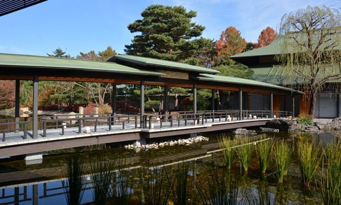 京都迎賓館庭園的廊橋在秋天：有屋頂的木造廊橋跨過池塘，後方松樹旁的楓樹已經轉紅，池邊長著一叢叢蘆葦，右側是垂柳與館舍