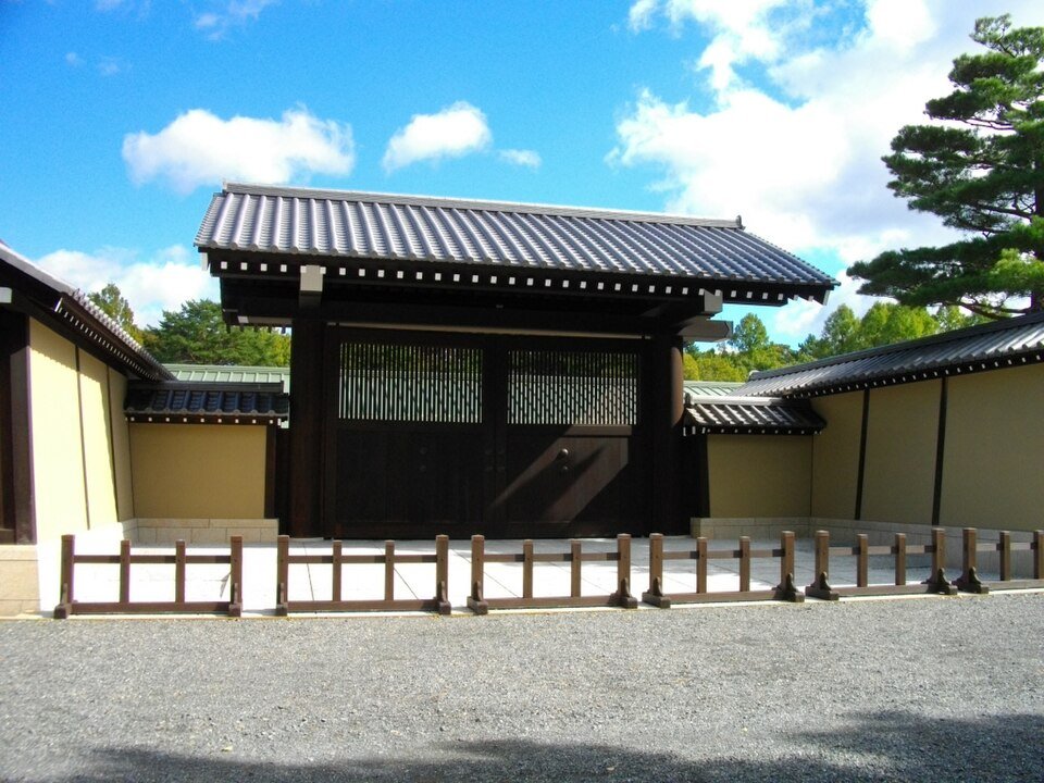 京都迎賓館的正門：深褐色木門扉、黑瓦屋頂的門，左右接著米黃色的圍牆，門前有一排低矮的木柵，地上是京都御苑的碎石