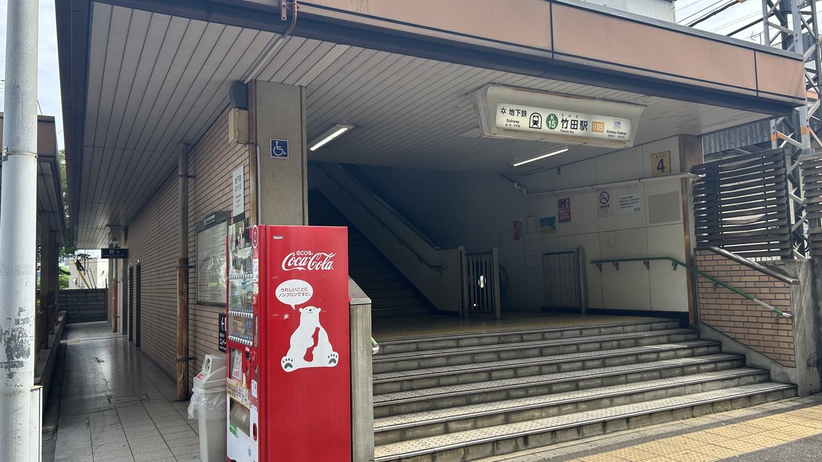 竹田站 4 號出入口:磚牆與米白色站房之間的樓梯口,簷下掛著「地下鉄 竹田駅」站名牌,右側牆上有黃底的「出入口 4」號牌,左前方是一台紅色自動販賣機