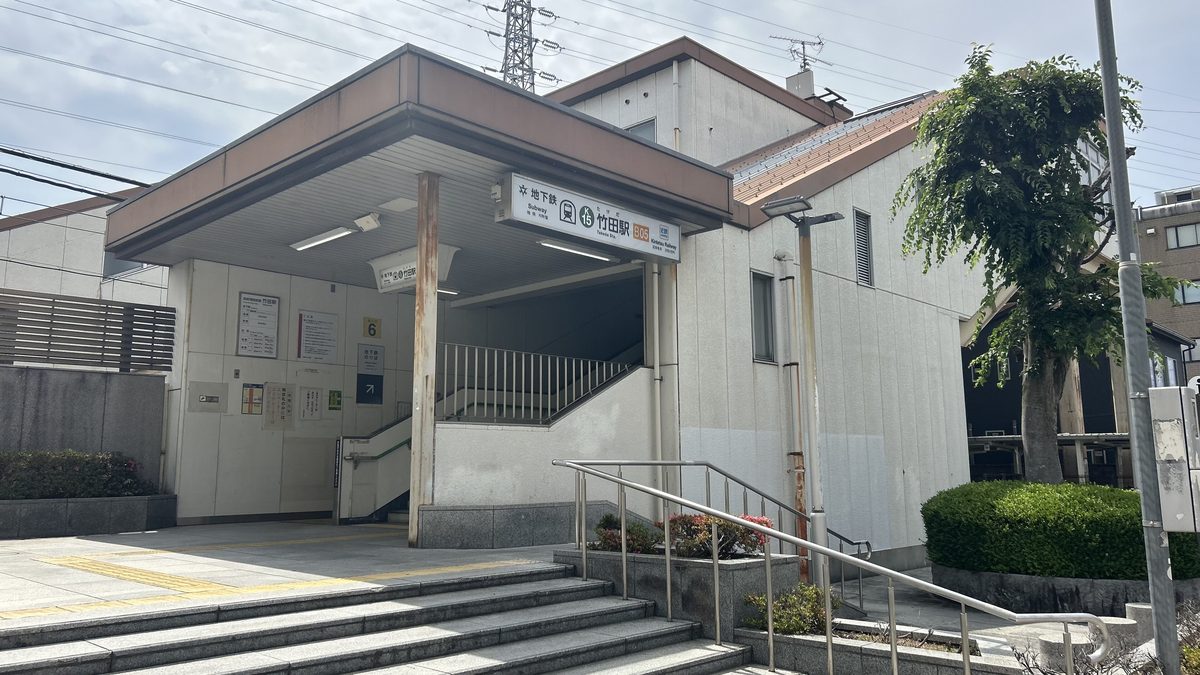 竹田站 6 號出入口的外觀:米白色站房下方的樓梯口,簷下掛著「地下鉄 竹田駅」站名牌,牆上有一塊黃底黑字的「6」號牌