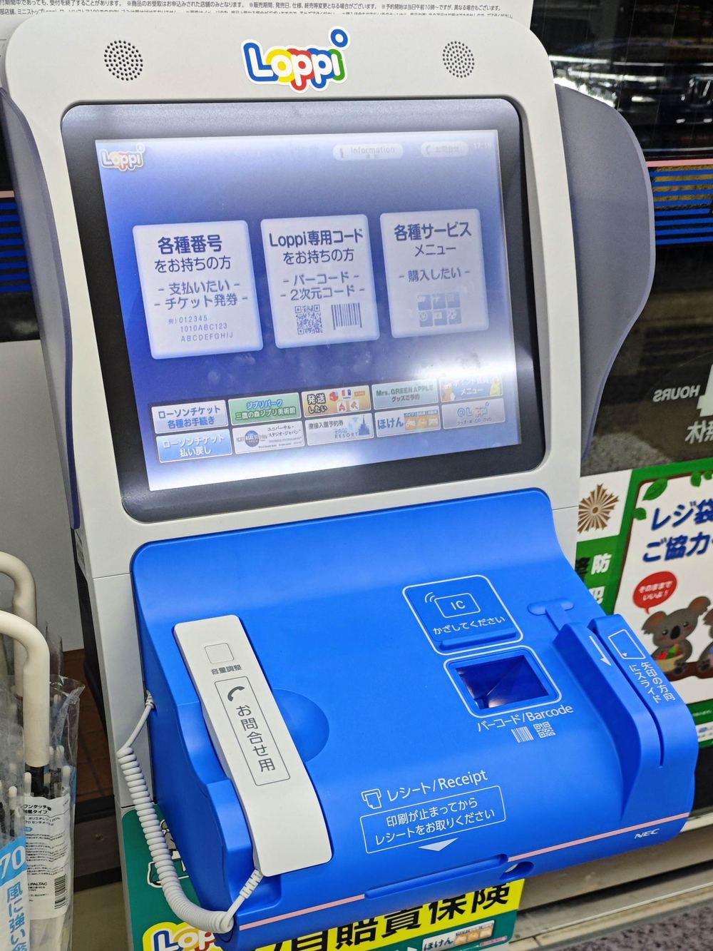 A white and blue Loppi terminal inside a Lawson store: a touchscreen on top showing three home-screen buttons for number holders, Loppi code holders and the services menu; below it a blue panel with an IC card pad, a barcode window, a coiled handset on the left labelled for enquiries, and a front edge printed with receipt instructions
