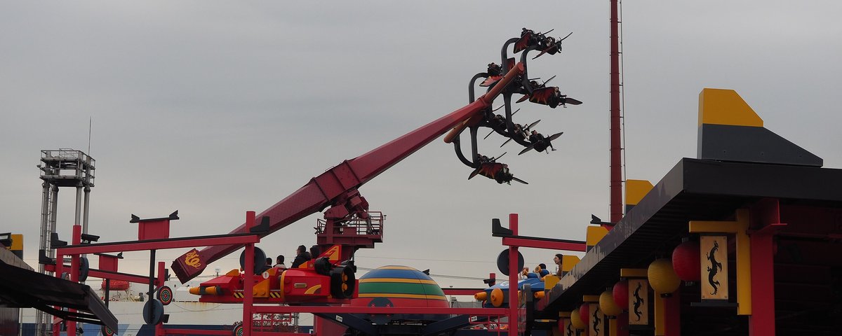 Flying NINJAGO:一根紅色長臂斜伸向陰天的天空,末端掛著一圈黑紅色的座椅,乘客被甩在半空中;下方是紅色鋼架、彩色條紋的圓頂和紅黑色的亭子,亭簷掛著紅黃燈籠與龍紋布條