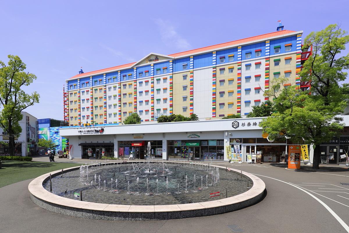 從メイカーズ ピア的圓形噴水池看樂高樂園飯店:前景是一座石砌圓形水池,中間噴著一圈細水柱;後面一排白色平房商店,招牌上有杉養蜂園等店名;再後面是紅色屋頂、牆面拼成彩色積木條紋的八層飯店,晴天