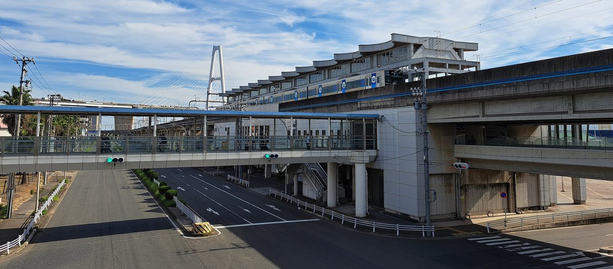 金城ふ頭站與站前天橋:右邊是灰白色的高架站房,屋頂是一節節波浪狀的屋簷,月台上停著一列白藍色的青波線電車;站房左側伸出一條藍色屋頂的有頂天橋,橫跨下方空曠的四線道路,橋上有幾位行人;遠方是白色的斜張橋塔,藍天白雲