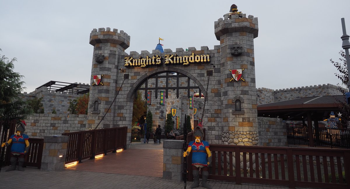 騎士王國的入口:灰色石磚造型的城門,兩座方塔中間掛著金色「Knight's Kingdom」招牌,塔身有紅底獅子盾徽,拱門下是鐵柵與彩色旗幟,前面是一座木欄杆小橋,橋兩側各站一尊藍衣騎士的積木像,陰天傍晚