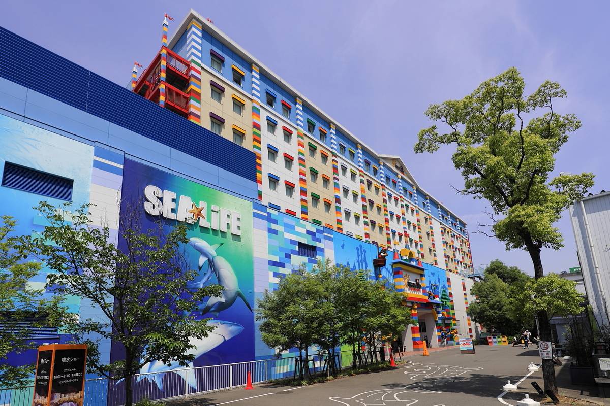 SEA LIFE 名古屋與樂高樂園飯店的外觀:左邊低層是藍色外牆、印著「SEA LIFE」字樣與兩條鯊魚的大幅壁畫,後面接著八層樓高、窗框塗成紅黃藍綠積木色的飯店,右邊一棵大樹,前面是畫著海豚線條的車道,晴天