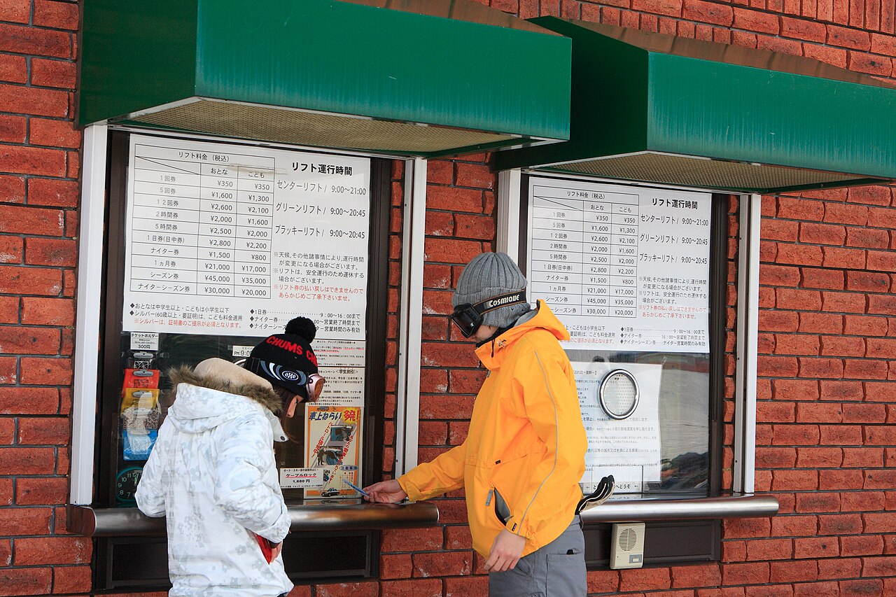 Lift ticket windows at a Japanese ski resort: two green-awninged hatches in a brick wall, fare boards taped to the glass with adult and child columns, two customers buying at the counter