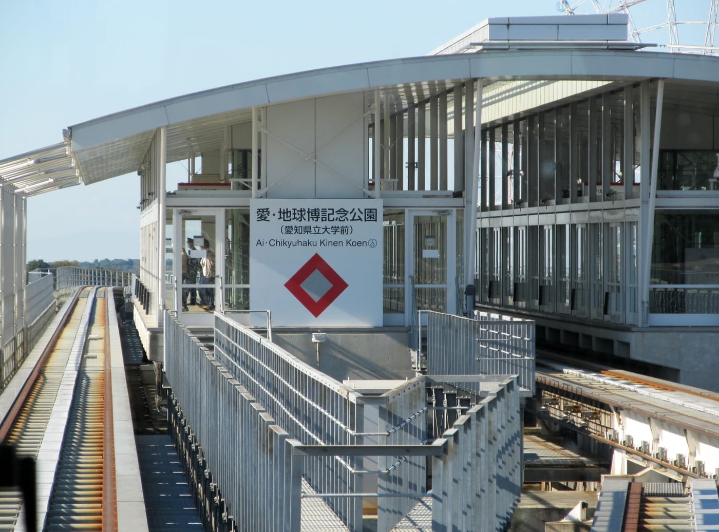 The elevated platform at Expo Memorial Park station on the Linimo maglev, with a white nameboard reading Ai・Chikyuhaku Kinen Koen and a red diamond emblem, and an empty guideway running away to the left