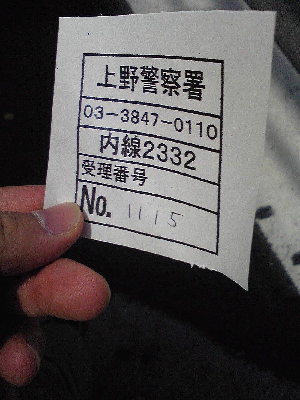 A hand holding a small torn slip of white paper printed with a boxed table: bold Japanese reading Ueno Police Station, the phone number 03-3847-0110, extension 2332, a row labelled reception number, and a final box marked No. with 1115 written in by hand