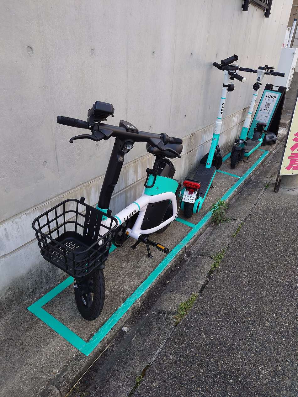A narrow pavement beside a concrete wall with individual parking boxes marked out on the ground in turquoise tape; nearest the camera sits a white-and-turquoise shared machine with a saddle and a black wire basket, behind it an electric kick scooter carrying a small number plate and red rear lamp, then two more scooters, and at the far end an A-frame board printed with a operator logo and a QR code