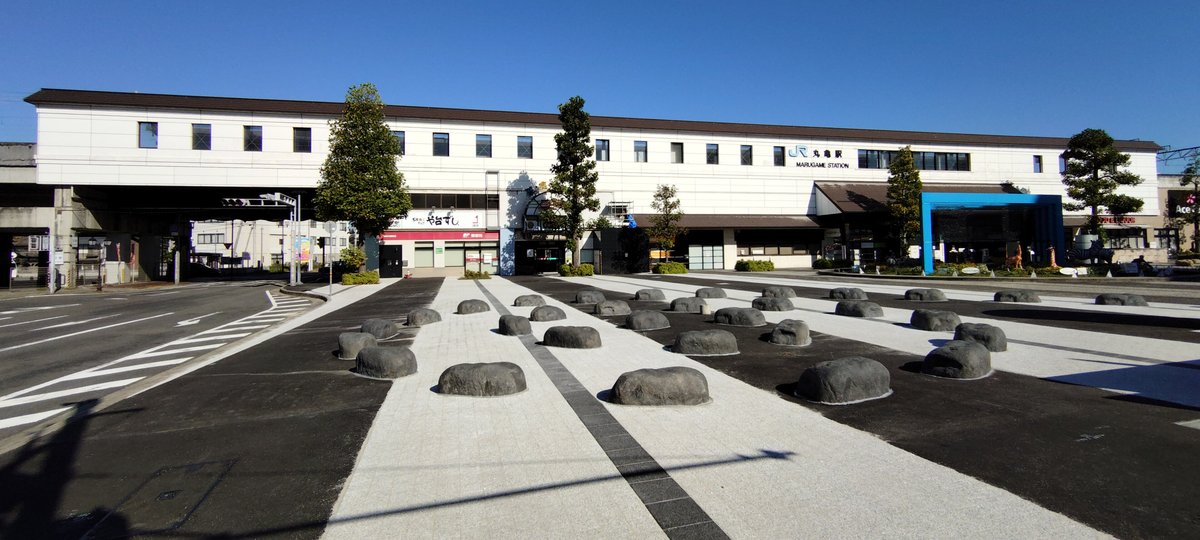 JR 丸龜站南口:一棟白色長條的高架站舍,牆上有 JR 標誌與「丸亀駅 MARUGAME STATION」字樣,右側入口是藍色框的玻璃門,站前廣場鋪著黑白條紋地磚,上面散放著一顆顆灰色的圓石