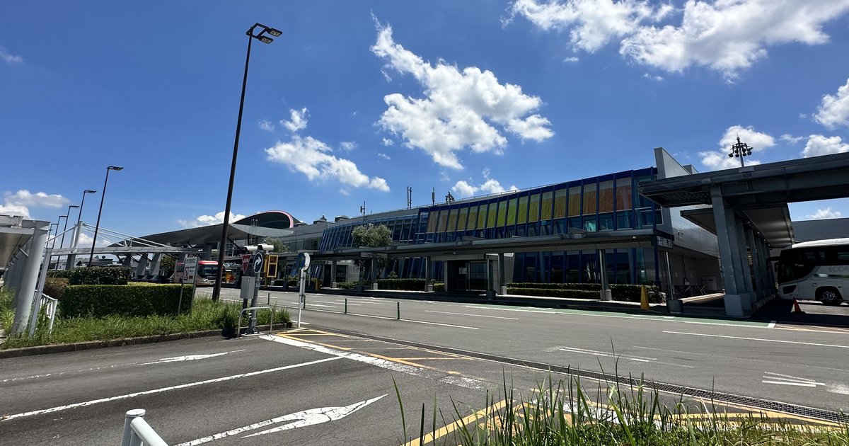 高松機場航廈正面:藍色與黃色玻璃帷幕的兩層建築,前方是寬闊的車道與白色箭頭標線,右邊有一排灰色柱子撐起的遮雨棚,棚下停著一輛白色巴士