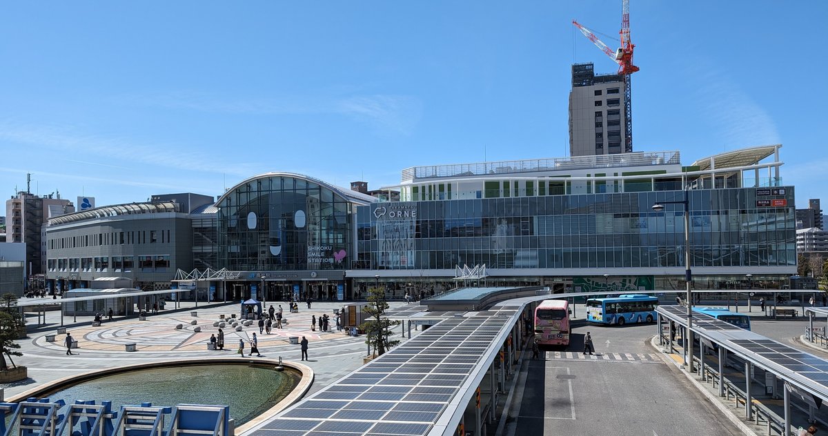 從天橋上看 JR 高松站:左邊是半圓拱形的玻璃站舍,上面寫著「SHIKOKU SMILE STATION」,右邊連著一棟玻璃帷幕的商業大樓「TAKAMATSU ORNE」,前方是鋪著圓形圖案的站前廣場,右側是有太陽能板頂棚的公車乘車處