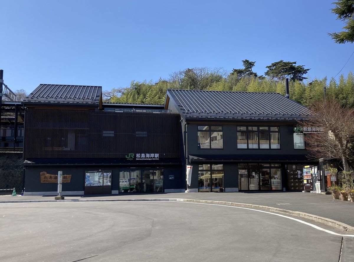 JR 松島海岸站的站房：兩棟相連、黑灰色外牆配深色斜屋頂的和風建築，左棟正面是木格柵外牆，招牌寫著 JR 標誌和「松島海岸駅 Matsushimakaigan Station」，一樓是整排玻璃門窗，站前是空曠的柏油廣場，背後山坡上長著松樹與竹林