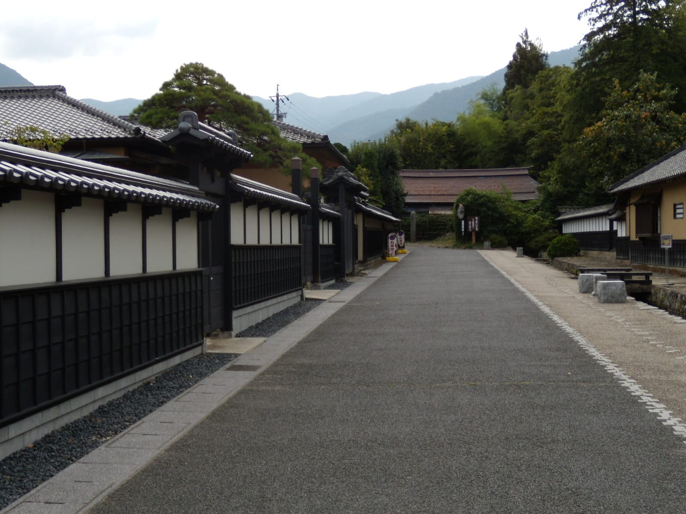 A former samurai-quarter street in Matsushiro: a long white wall with black wooden lattice panels running down the left side, a wide paved roadway in the centre, stone benches and a water channel on the right, a dark-roofed gatehouse closing the far end, and layered mountain ridges beyond