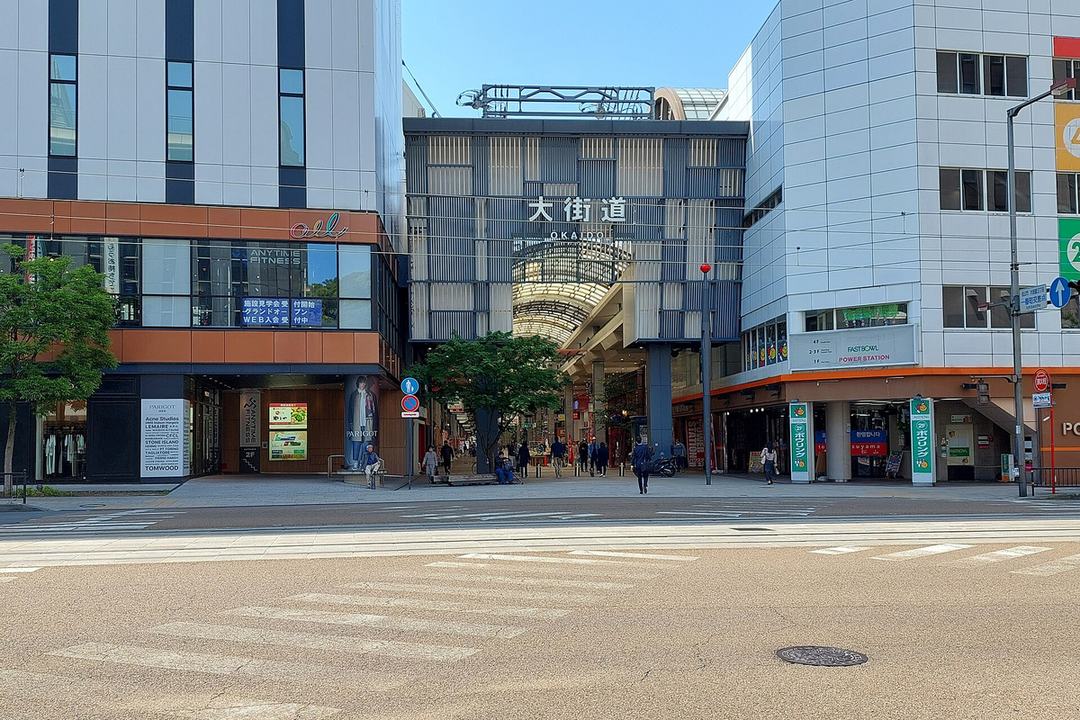 隔著有電車軌道的寬馬路看大街道商店街的入口:兩棟大樓之間的拱廊入口上方掛著「大街道」三個大字,拱廊裡往深處延伸,入口前有幾個行人