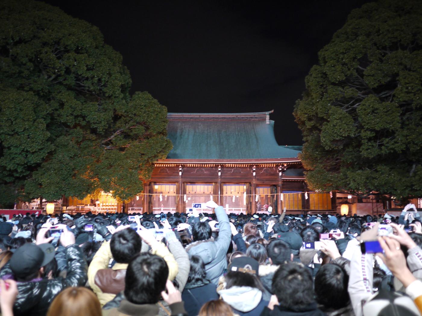 Dense hatsumode crowd in front of a shrine worship hall, visitors holding up phones