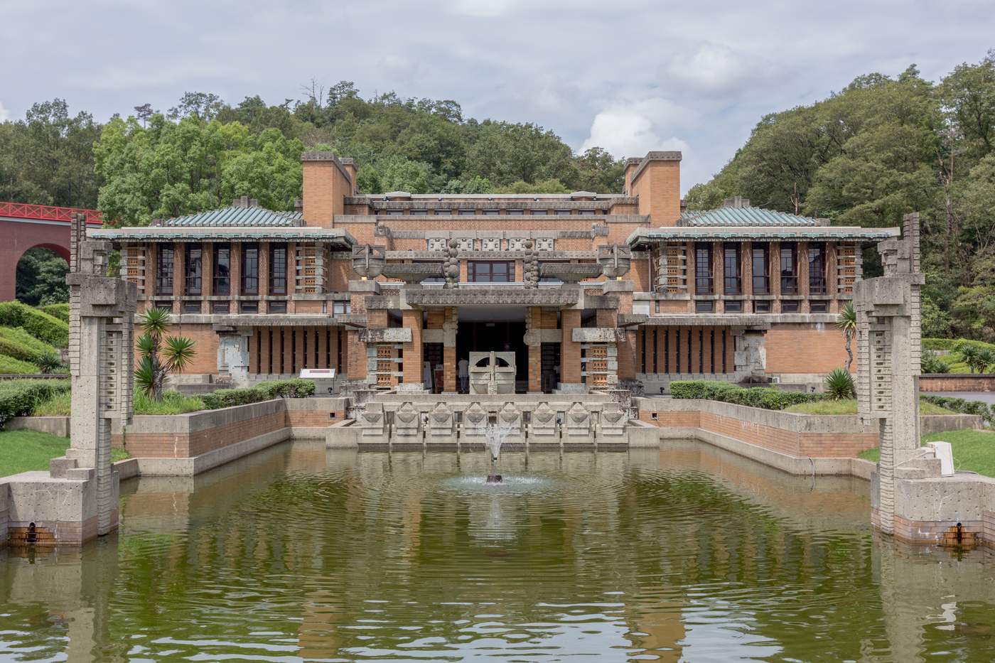 Front elevation of the Imperial Hotel entrance hall, a Registered Tangible Cultural Property: a symmetrical facade of brick banded with pale carved stone spreading into two wings, above a rectangular reflecting pool with a single fountain jet, a carved stone pier standing at each end of the pool