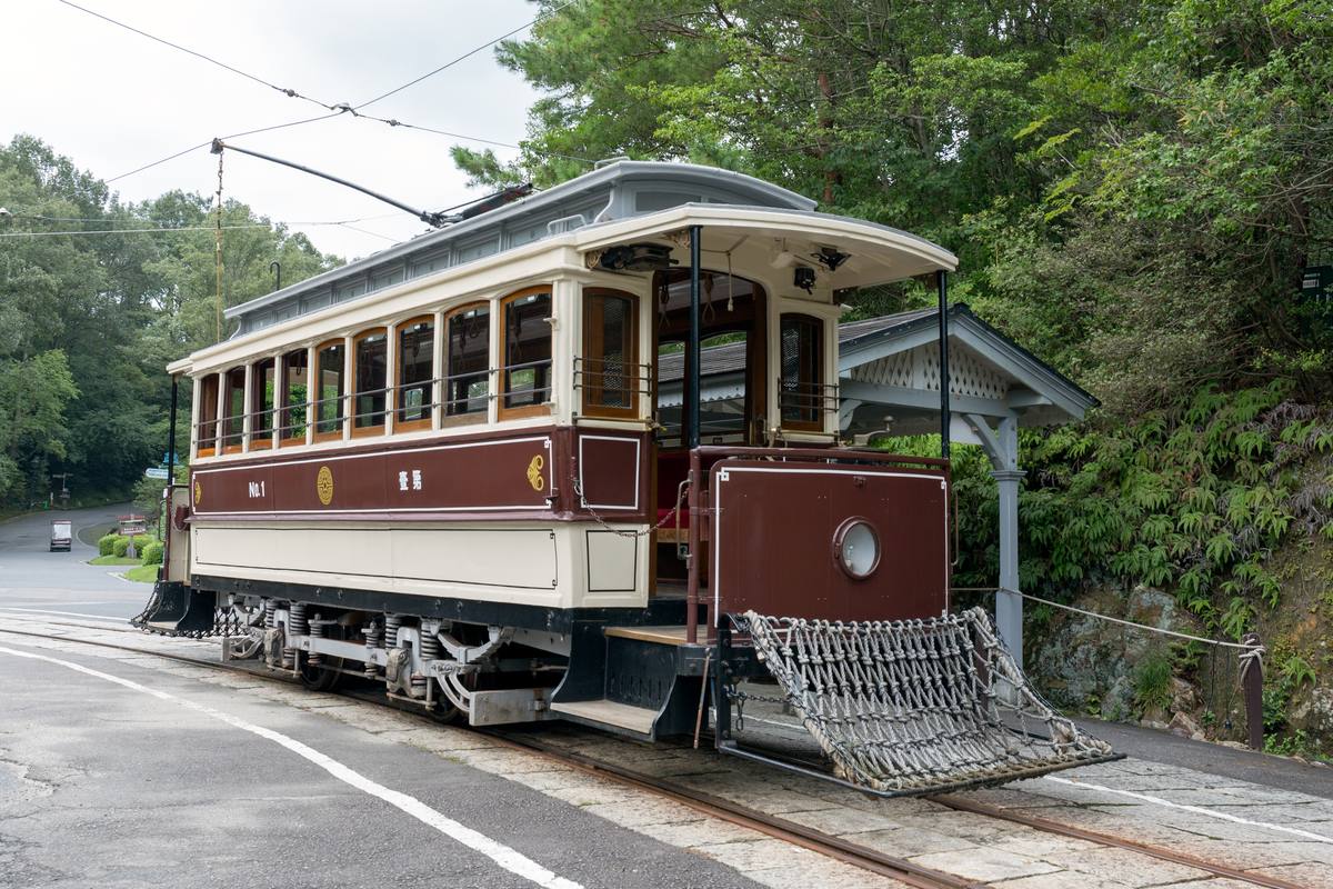 The Kyoto tram at Meiji-mura standing beside a timber waiting shelter, gold lettering reading No. 1 on its deep red body, a woven mesh fender below the front platform and the trolley pole reaching up to the overhead wire