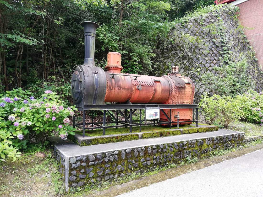 A rust-red steam locomotive boiler laid on an outdoor steel stand, black smokebox and chimney at the near end, an interpretation board fixed to the front of the stand and hydrangeas in flower on both sides
