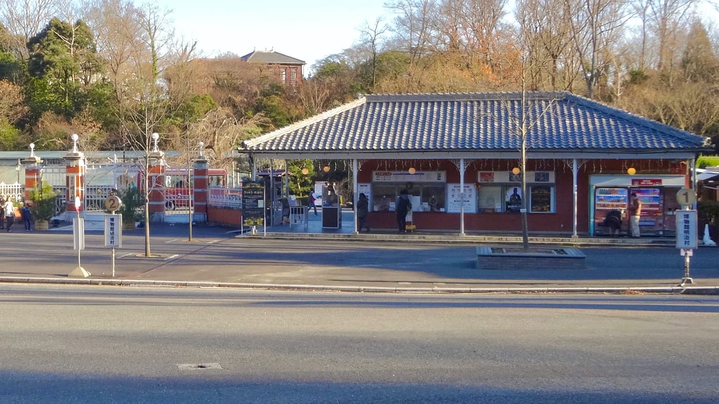 The bus turning circle in front of the Meiji-mura main gate: on the right a vermilion timber ticket office under a dark tiled roof with a few visitors at the windows and vending machines alongside; on the left a set of brick-red gate piers with white iron gates; in the foreground two slim bus-stop poles reading 博物館明治村, numbered 1 and 2; bare winter trees under a clear sky