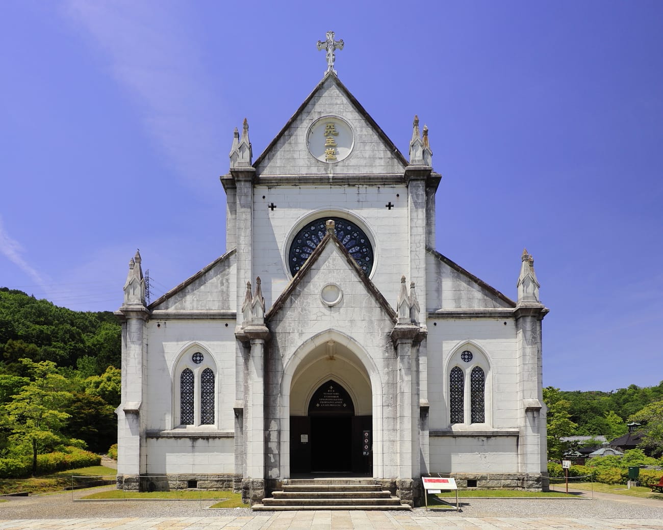 The facade of St Francis Xavier's Cathedral at Meiji-mura: a symmetrical grey stone Gothic Revival front with a silver cross on the gable apex, a circular medallion in the gable carrying the gilded characters 天主堂, a stained-glass rose window below it, a Latin inscription over the pointed-arch doorway, lancet windows and small pinnacles either side, green hills behind and a blue sky