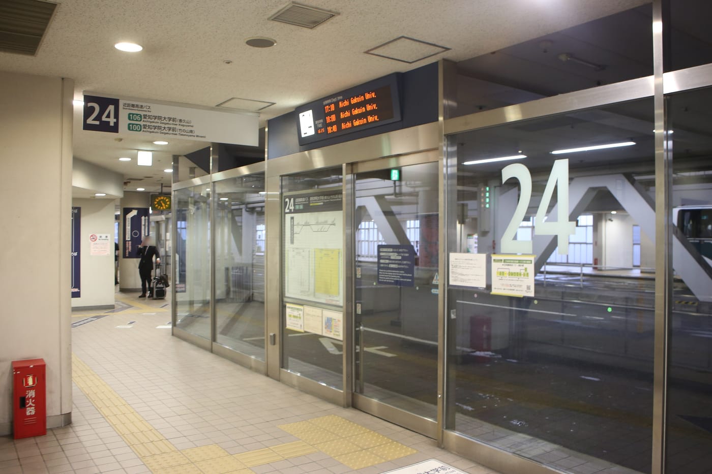 Bay 24 on the fourth floor of the Meitetsu Bus Center: a wall of full-height glass doors with a large number 24 printed on them, a blue sign above reading bay 24 short-distance highway buses with routes 106 and 100 to Aichi Gakuin University, and a departure board listing 17:10, 18:00 and 18:40