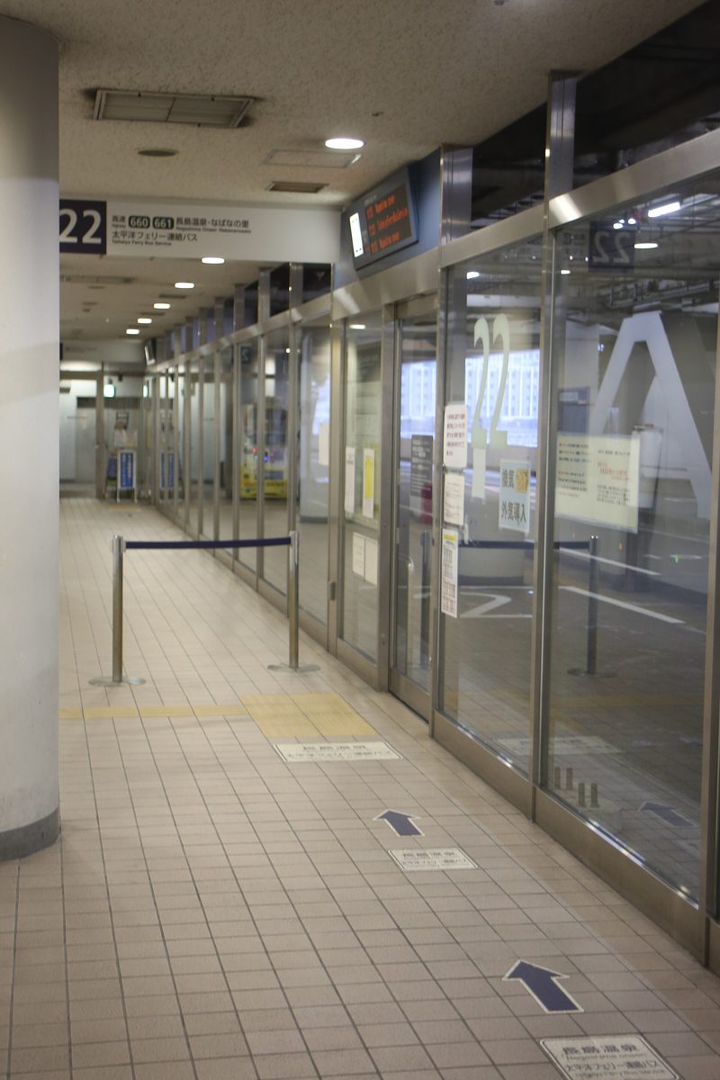Indoor waiting corridor at Meitetsu Bus Center: an overhead sign reads 22, Highway 660 661, Nagashima Onsen / Nabananosato and Taiheiyo Ferry Bus Service; glass doors and a departure board on the right, arrow stickers on the floor tiles