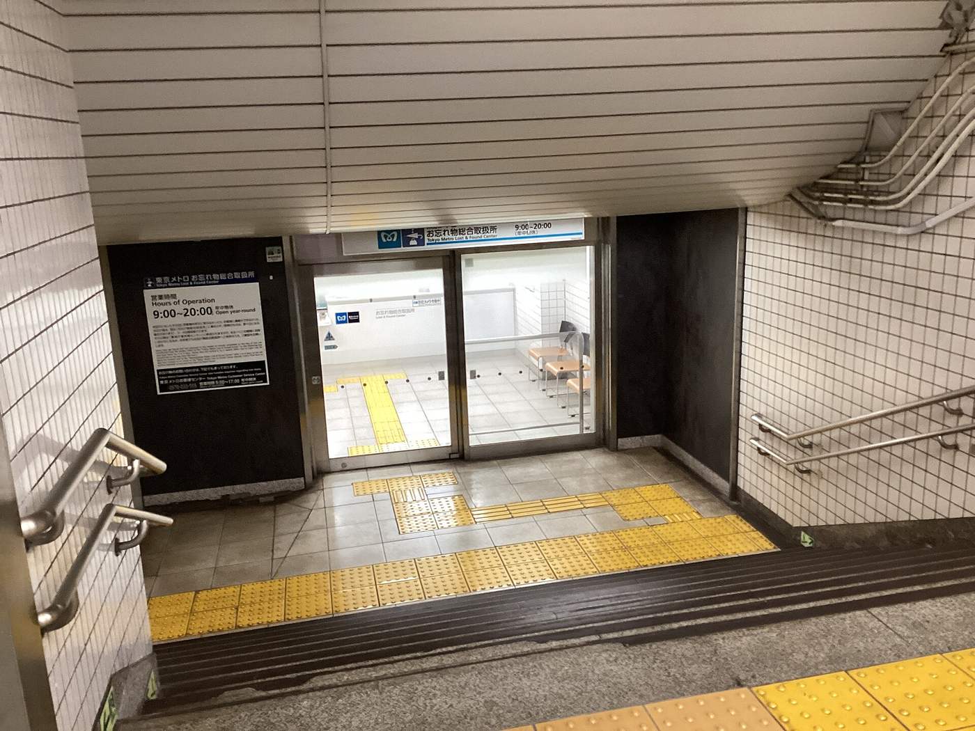 Looking down a staircase to the end of a subway passage, where a blue and white sign above glass automatic doors reads Owasuremono Sogo Toriatsukaijo, Tokyo Metro Lost & Found Center, 9:00-20:00, open year-round; a black notice board beside the doors repeats the hours, yellow tactile paving crosses the floor, and orange waiting chairs are visible through the glass