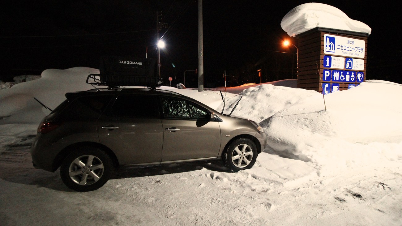 A roadside-station car park at night: an SUV with a roof cargo box parked on packed snow, a silver insulating screen behind its windscreen and the wipers propped up, beside the snow-capped pylon sign of Michi-no-Eki Niseko View Plaza with its parking and toilet pictograms